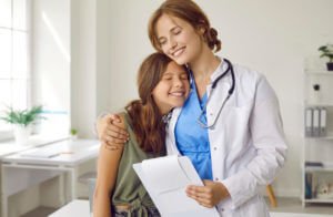 Happy pediatrician standing in medical office, hugging child patient and smiling.