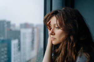 Young woman with brown hair stares through a rain-streaked window in a city apartment, reflecting anxiety and depression.