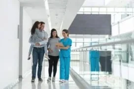 A mother carries her toddler while walking beside a nurse in a bright hospital corridor