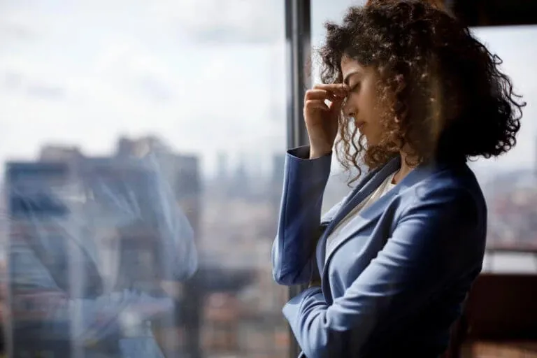 A woman pauses at a window, holding her forehead while managing feelings of stress and anxiety