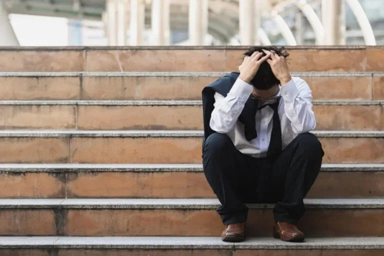 A stressed businessman sits on outdoor stairs with his head in his hands, reflecting the work-related anxiety Abundance Health Care supports patients through