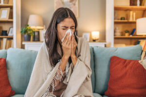 Woman suffering from a common cold, sitting on her couch, covered with a blanket, and blowing her nose.